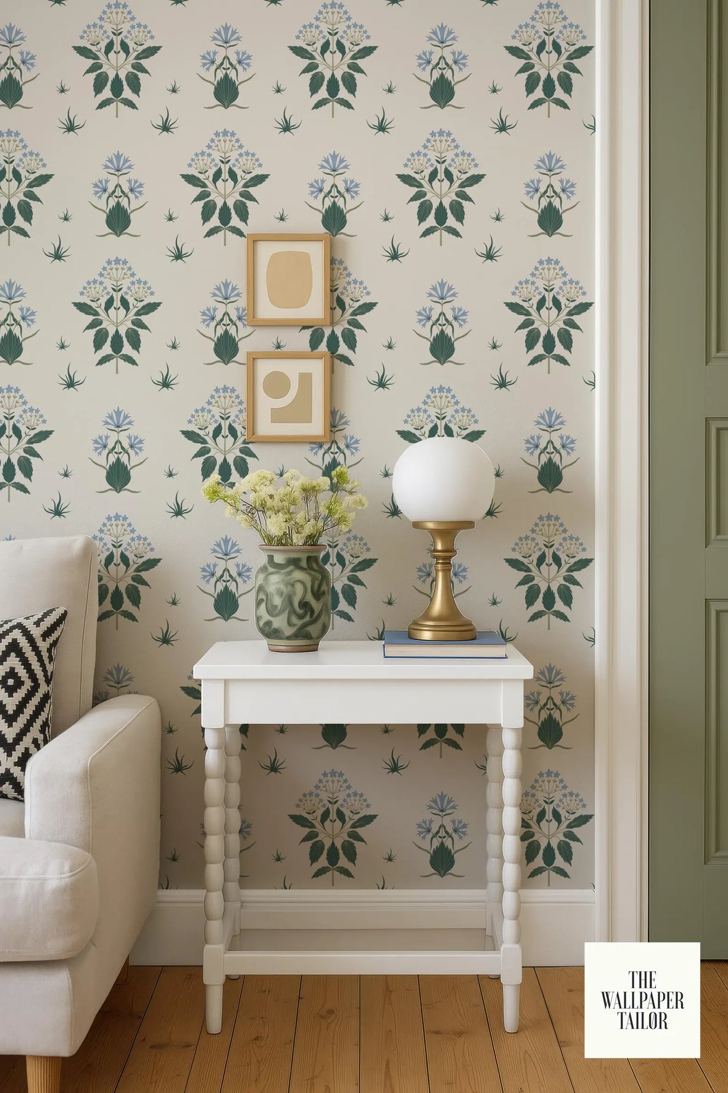 a cozy living room with a white sofa, a white side table with a lamp and a vase of flowers, and a patterned wallpaper.