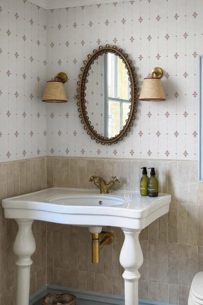 a bathroom with a white pedestal sink, a large oval mirror with a gold frame, and two wall sconces with beige shades. The walls are covered in a patterned wallpaper, and the overall color scheme is neutral, with a touch of gold accents.