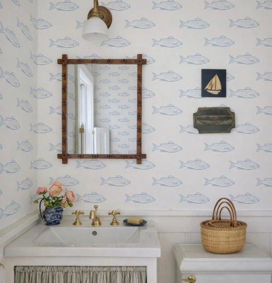 a bathroom with a white sink, a mirror, and a wooden frame. The walls are covered in a wallpaper with blue fish designs, and there is a small boat picture on the wall. A basket is placed on the floor next to the sink, and a vase of flowers is on the countertop.