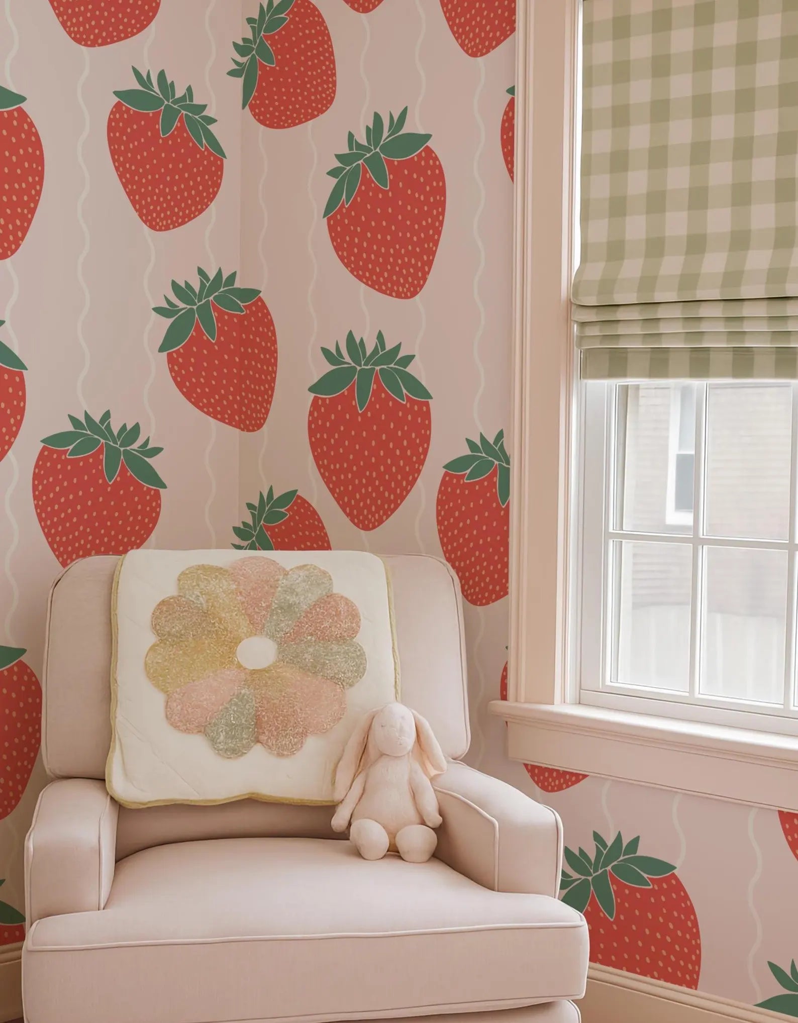 a white chair with a flower-shaped pillow on it, placed in front of a wall with a strawberry wallpaper. A stuffed bunny is sitting on the chair, and there is a window with a green and white checkered curtain in the background.
