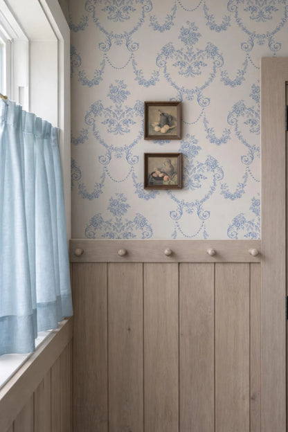 a cozy corner with a window, a wooden wall, and a wallpapered wall. There are two framed pictures hanging on the wall, and a curtain is partially drawn, allowing some light to enter the room.