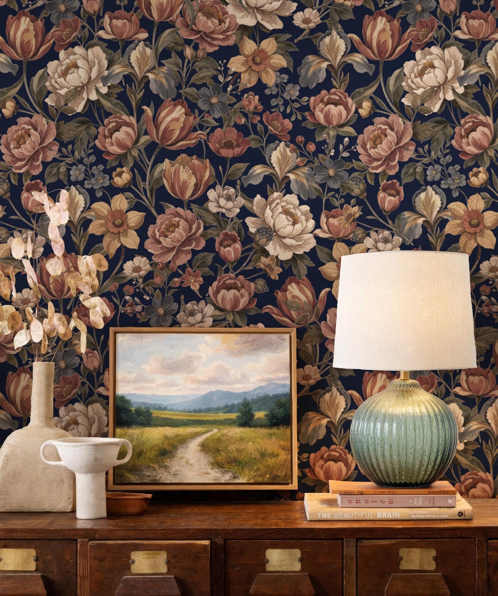 a wooden dresser with a painting of a countryside landscape, a lamp, and a vase on top. The dresser has a dark wood finish and is set against a wall with a floral wallpaper. The painting is framed and hangs on the wall, while the lamp and vase are placed on the dresser.