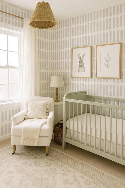 a serene and cozy nursery room with a white crib, a white armchair, and a lamp. The room has a light green color scheme, and the walls are adorned with a patterned wallpaper. There is a window in the room, allowing natural light to enter, and a wicker pendant light hanging above the crib. The room also features two framed pictures of a rabbit on the wall.