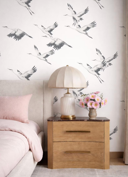 a bedroom setting with a bed, a wooden nightstand, and a lamp with a white shade. The wall behind the nightstand has a repeating pattern of black and white birds in flight.