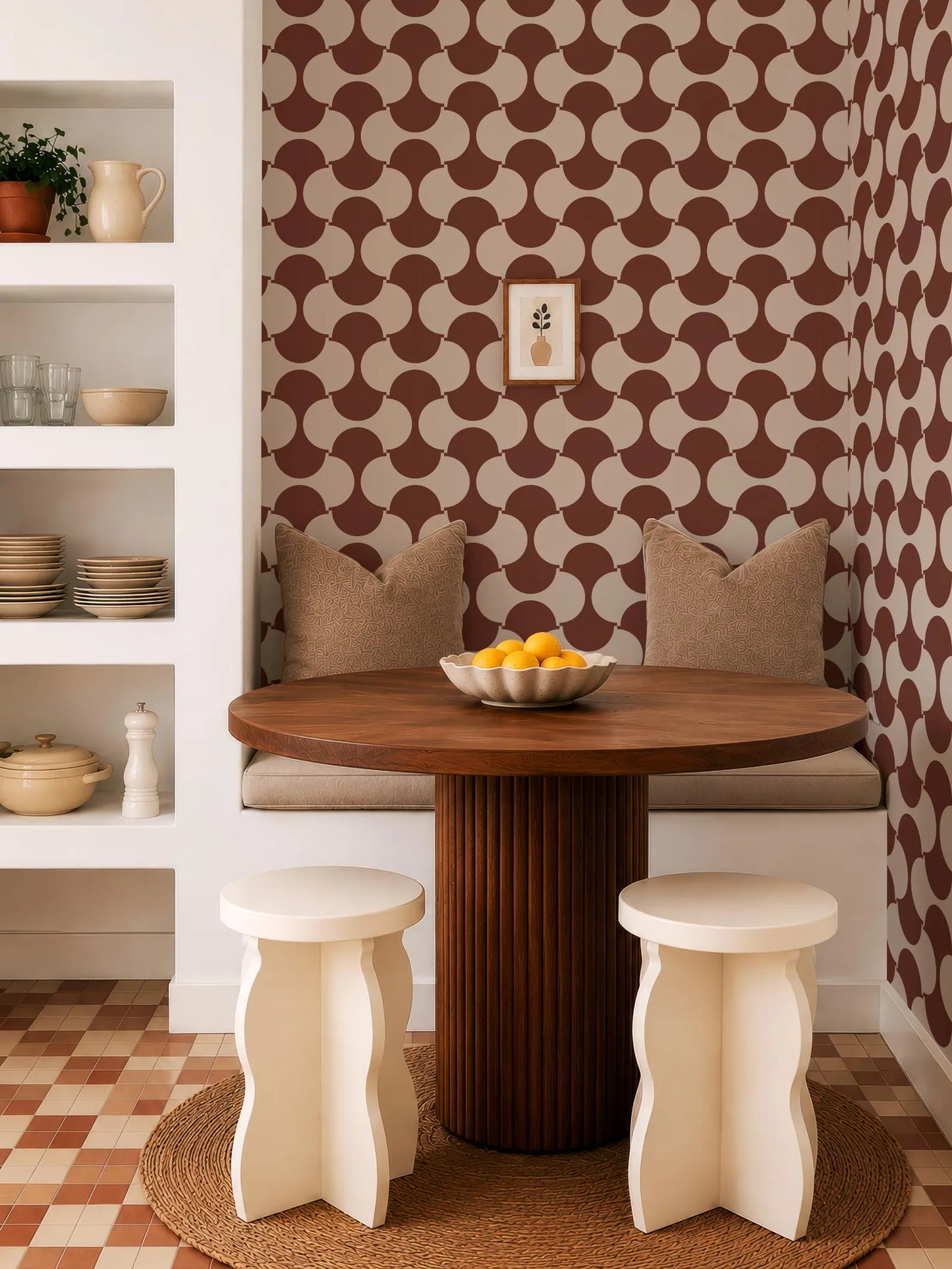 a modern dining area with a wooden table, two white stools, and a patterned wall behind the seating area. The table is set with a bowl of fruit, and there are decorative elements such as a vase and a plant in the room.