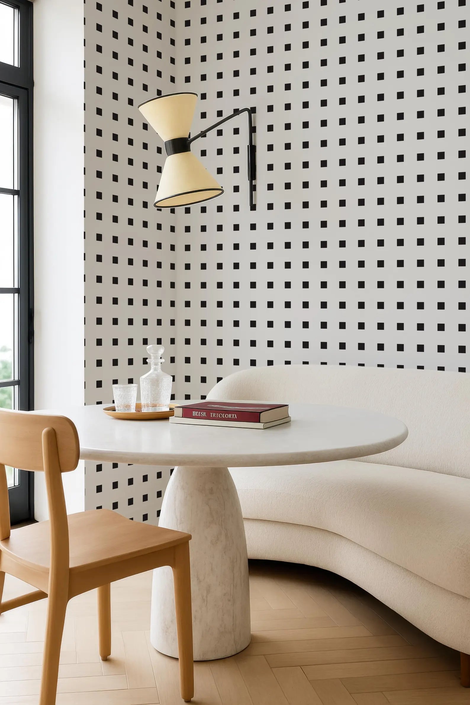 a modern living room with a white sofa, a round white table, and a wooden chair. The walls are covered in a black and white checkered pattern, and a lamp is hanging from the ceiling. On the table, there is a book and a bottle of wine.