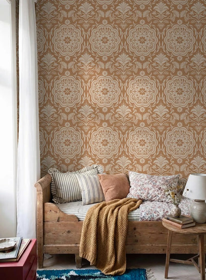 a cozy living room with a wooden bench, a striped pillow, a brown blanket, and a small table with a lamp and a vase of flowers. The walls are covered in a patterned wallpaper with a floral design in shades of brown and beige.