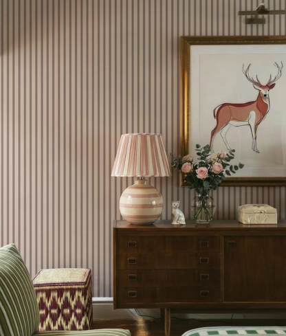 a cozy living room with a striped wallpaper, a wooden dresser, a lamp, a framed deer artwork, and a vase of flowers.