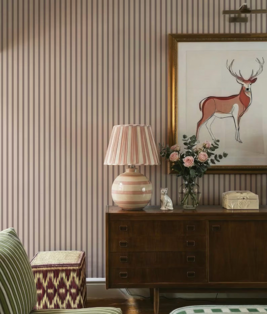 a cozy living room with a striped wallpaper, a wooden dresser, a lamp, a framed deer artwork, and a vase of flowers.
