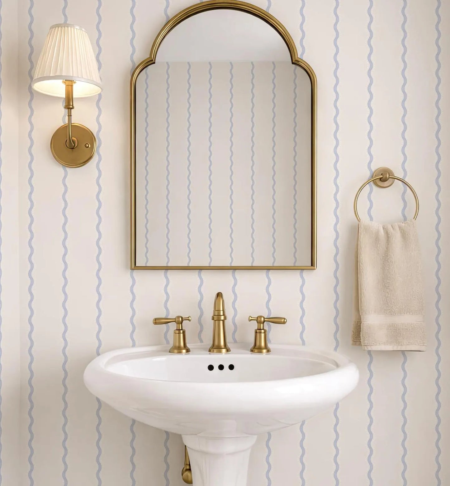 a bathroom with a white pedestal sink, a gold faucet, and a gold mirror above it. The walls are covered in blue and white striped wallpaper, and there is a towel rack with a towel hanging on it.