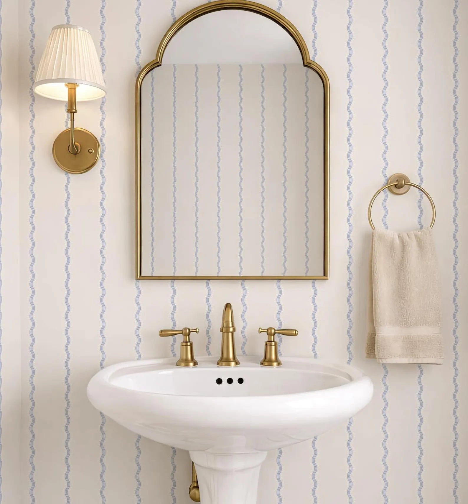 a bathroom with a white pedestal sink, a gold faucet, and a gold mirror above it. The walls are covered in blue and white striped wallpaper, and there is a towel rack with a towel hanging on it.