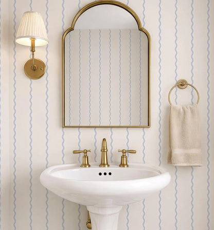 a bathroom with a white pedestal sink, a gold faucet, and a gold mirror above it. The walls are covered in blue and white striped wallpaper, and there is a towel rack with a towel hanging on it.