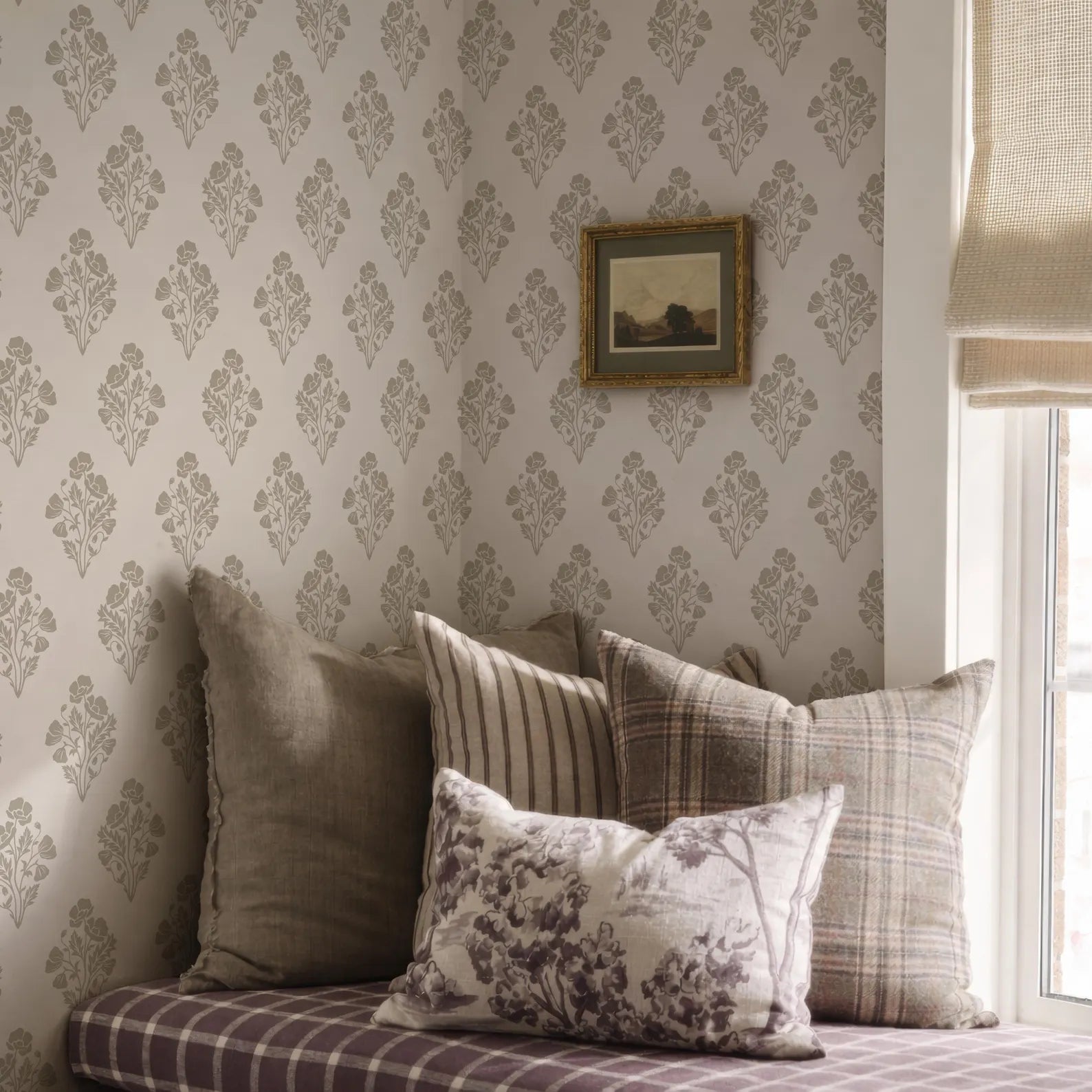 a cozy corner of a room with a plaid pillow, a striped pillow, and a floral pillow, all resting on a bed. The wall behind the pillows is covered in a patterned wallpaper, and there is a framed picture hanging on the wall.