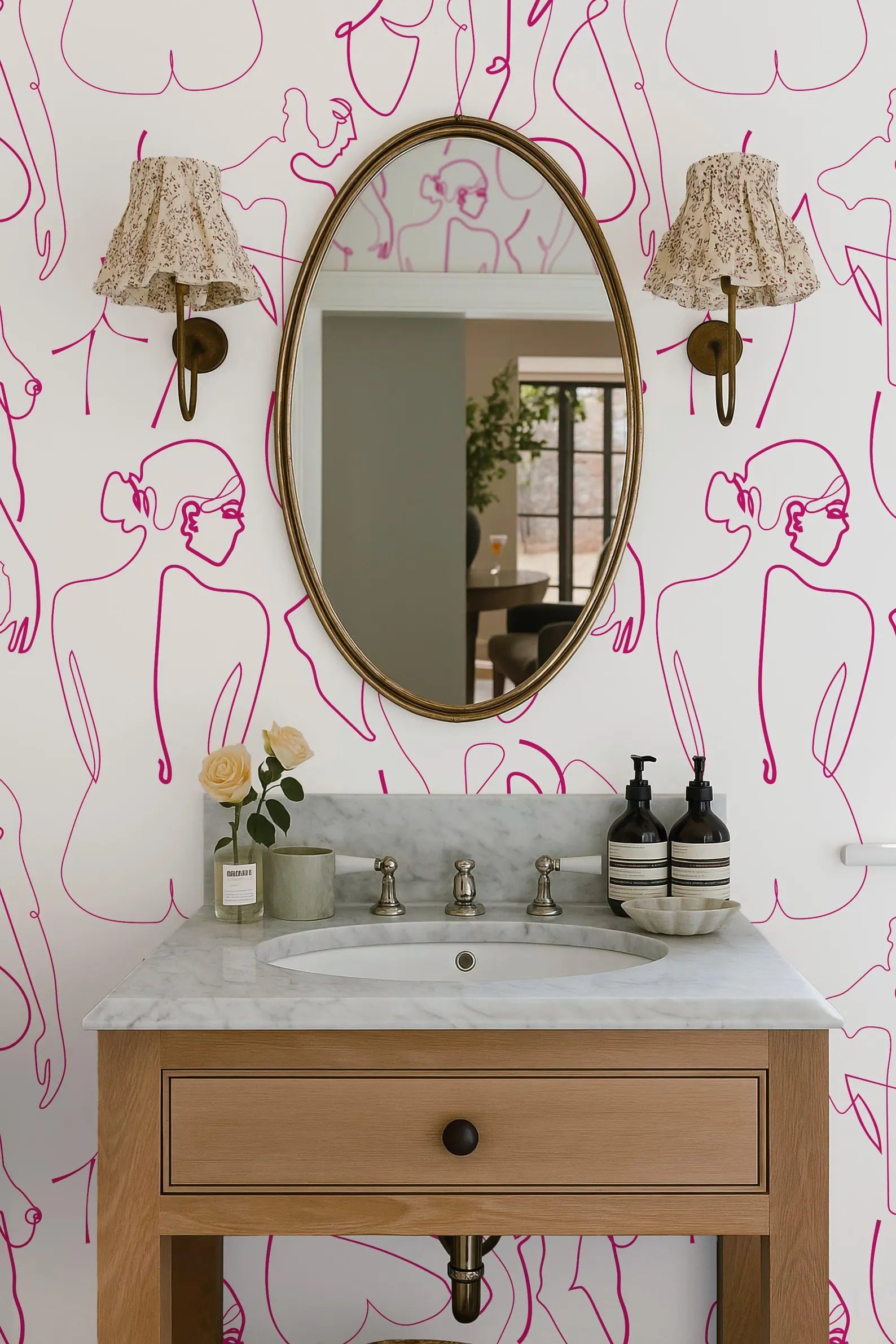 a bathroom vanity with a white marble countertop, a sink, and a mirror above it. The wall behind the vanity has a pink and white abstract pattern. There are two lamps on either side of the mirror, and a vase with a single rose is placed on the countertop.