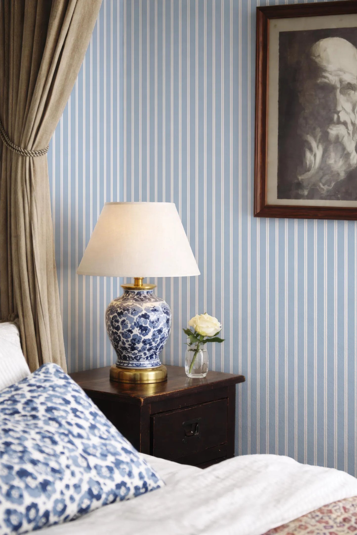 a bedroom with a blue and white striped wallpaper, a wooden nightstand with a lamp and a vase of flowers, and a framed picture on the wall.