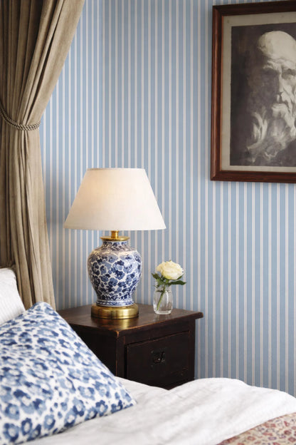 a bedroom with a blue and white striped wallpaper, a wooden nightstand with a lamp and a vase of flowers, and a framed picture on the wall.