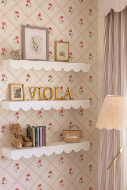 a room with a floral wallpaper, white shelves, and a lamp. On the shelves, there are several framed pictures, a teddy bear, and a basket with the name "Leona" written on it.