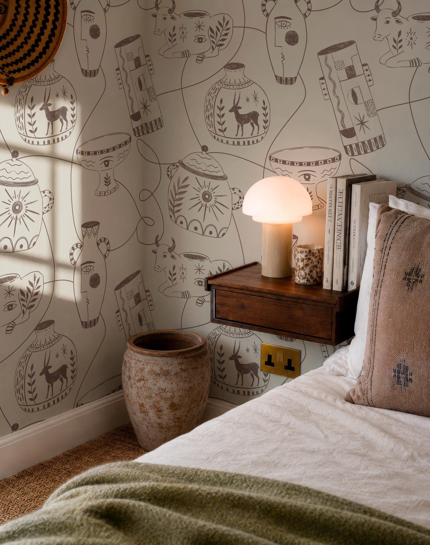 a cozy bedroom scene with a bed, a wooden nightstand, and a lamp. The wall behind the nightstand is decorated with a pattern of various objects, including vases, bowls, and other decorative items.