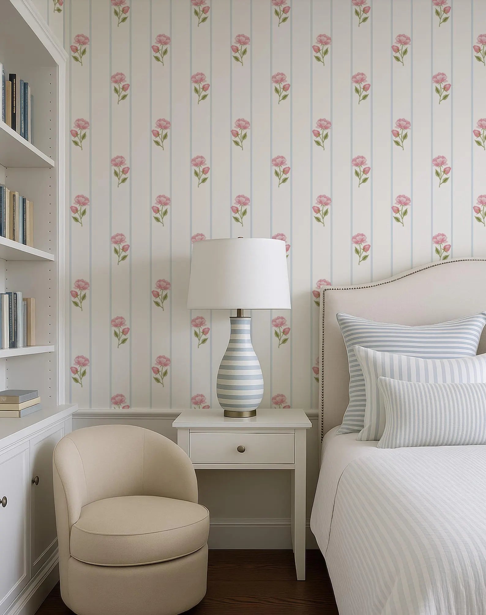 a cozy bedroom with a white bed, a white armchair, and a white nightstand with a lamp. The walls are covered in a floral wallpaper with pink flowers, and there are several bookshelves in the room.
