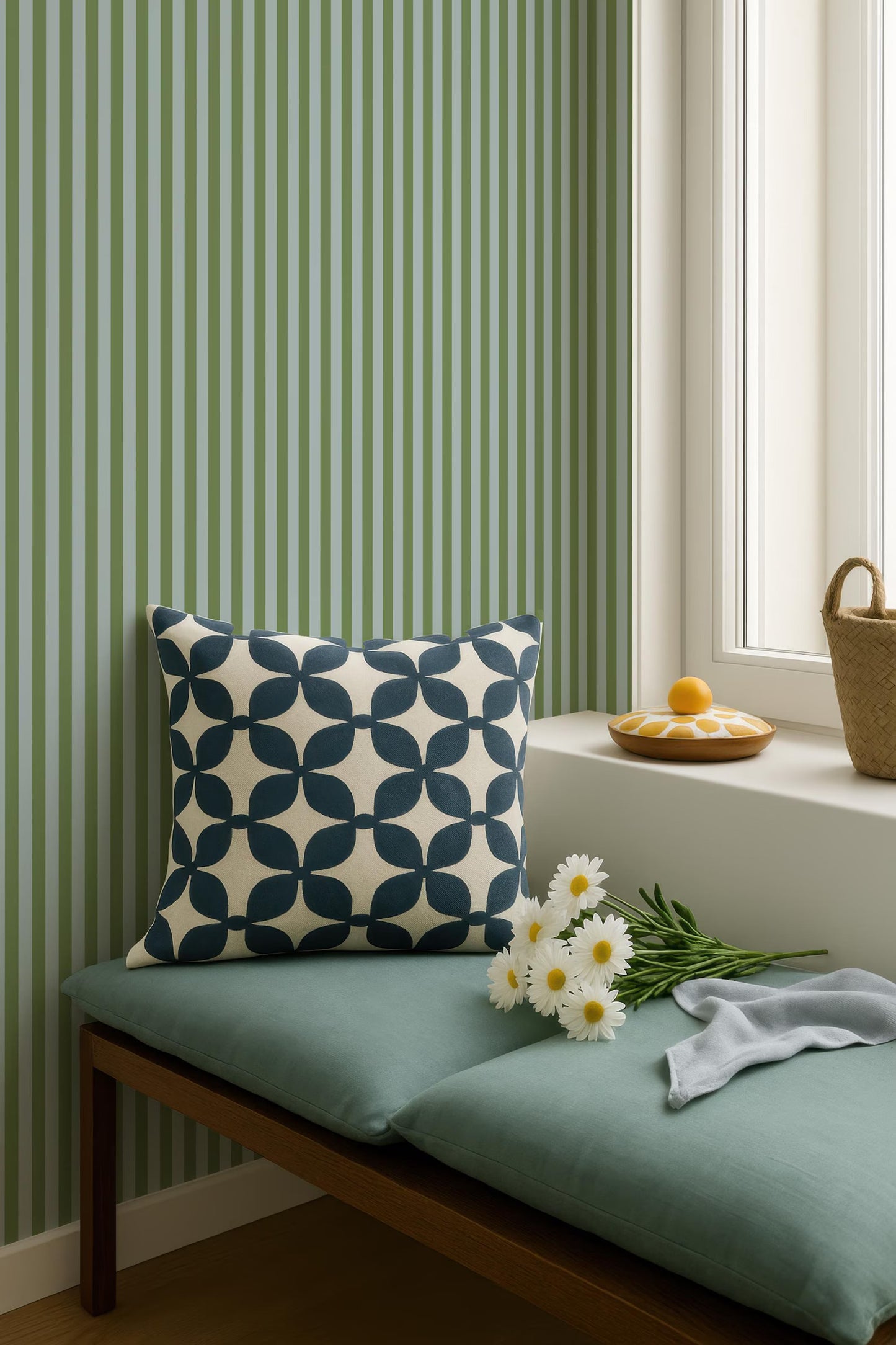 a cozy corner with a green and white striped wall, a blue and white patterned pillow, a basket of flowers, and a small wooden tray with an orange.