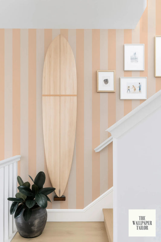 a wooden surfboard mounted on a wall, with a potted plant placed next to it. The wall has a striped pattern in shades of peach and white. There are also framed pictures hanging on the wall, adding to the overall decor.