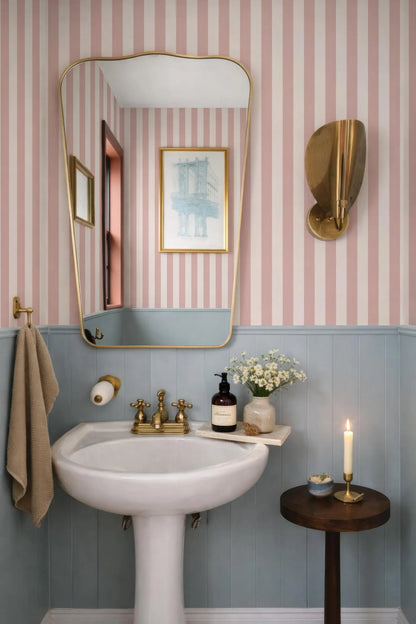a bathroom with a white pedestal sink, a mirror, and a small table with a candle. The walls are striped in pink and white, and there is a framed picture on the wall.