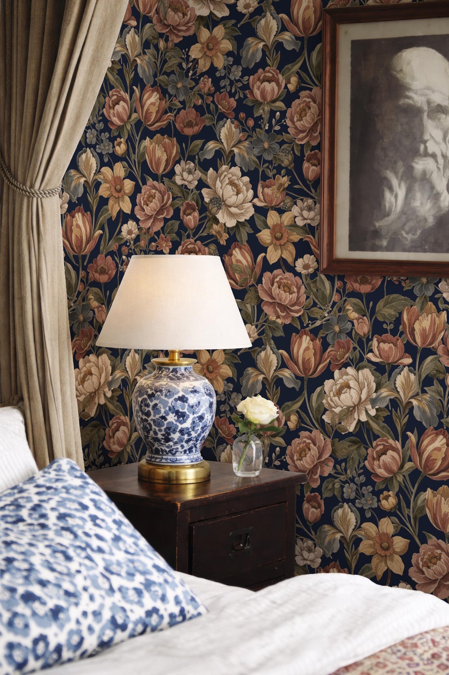 a bedroom with a floral wallpaper, a blue and white patterned pillow, a wooden nightstand with a lamp, and a framed picture on the wall.