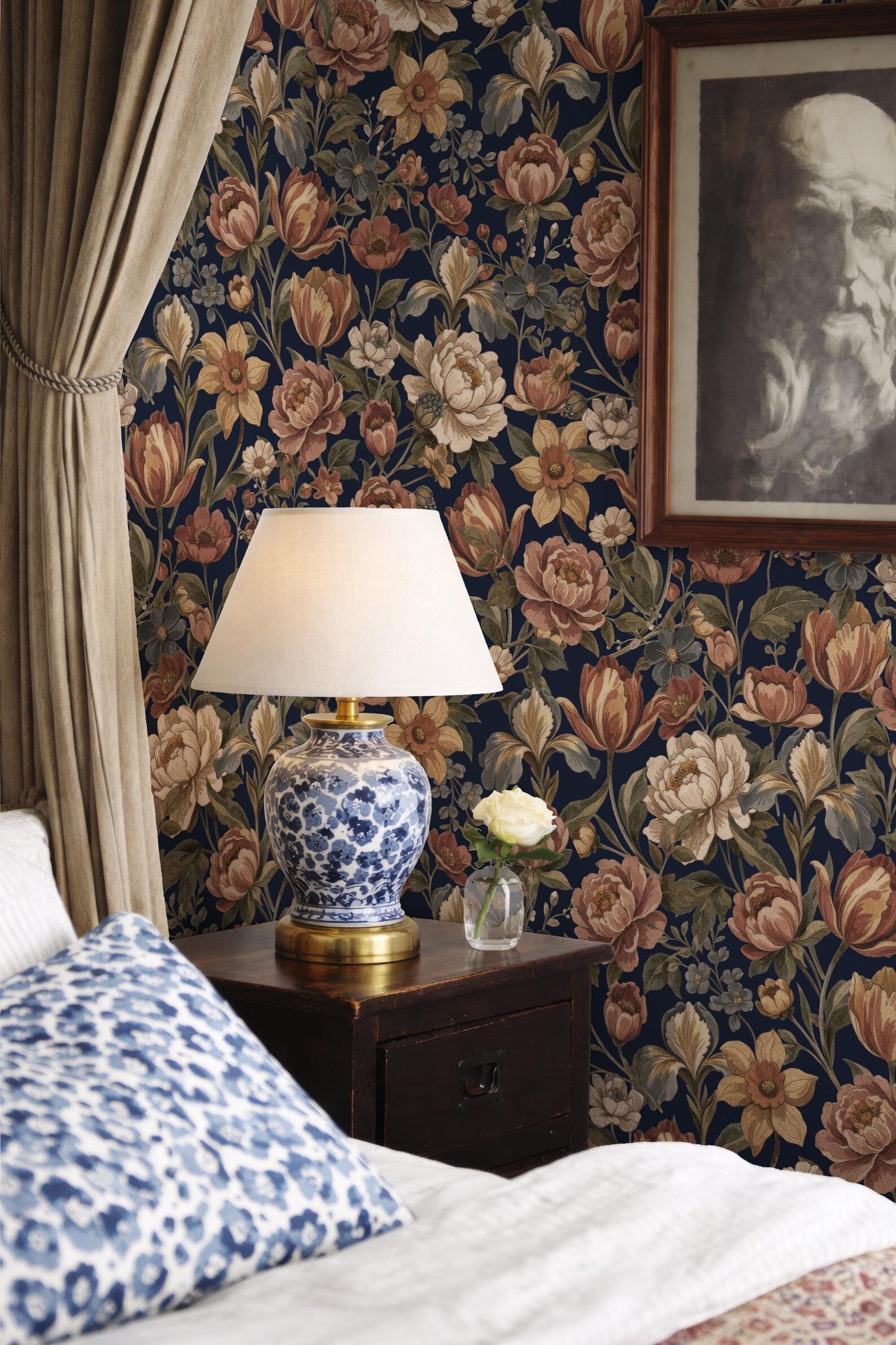 a bedroom with a floral wallpaper, a blue and white patterned pillow, a wooden nightstand with a lamp, and a framed picture on the wall.