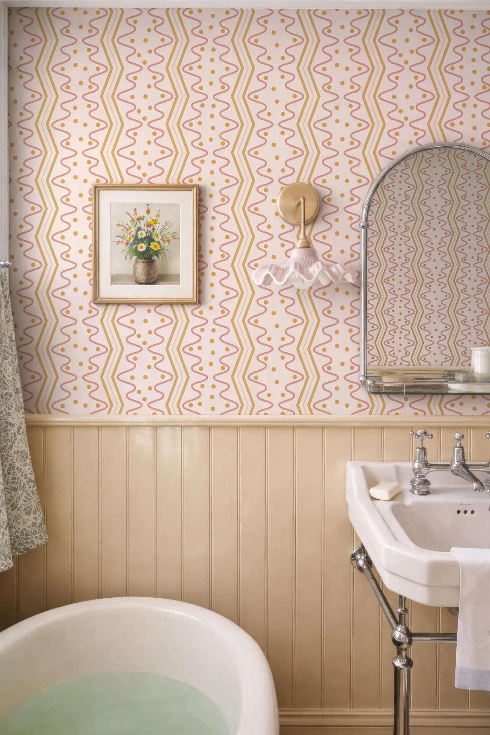 a bathroom with a white bathtub, a sink, and a mirror. The walls are covered in a patterned wallpaper, and there is a framed picture on the wall.