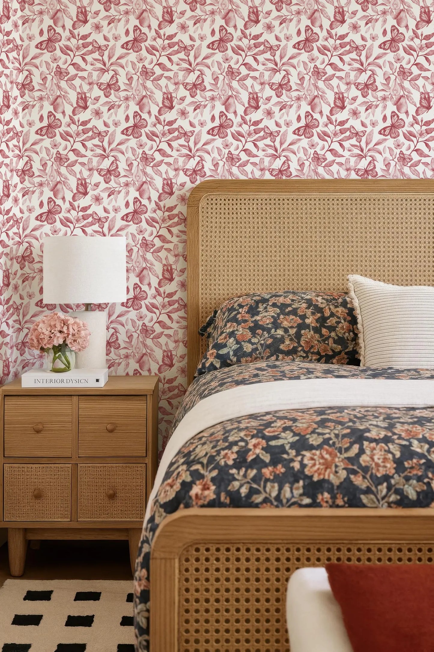 a bedroom with a floral bedspread, a wooden headboard, and a wooden nightstand with a lamp and a book. The walls are covered in a pink and white floral wallpaper, and the room has a cozy and inviting atmosphere.