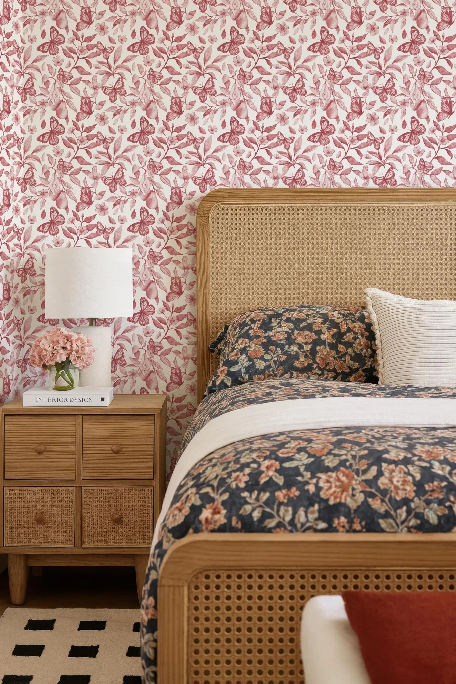 a bedroom with a floral bedspread, a wooden headboard, and a wooden nightstand with a lamp and a book. The walls are covered in a pink and white floral wallpaper, and the room has a cozy and inviting atmosphere.