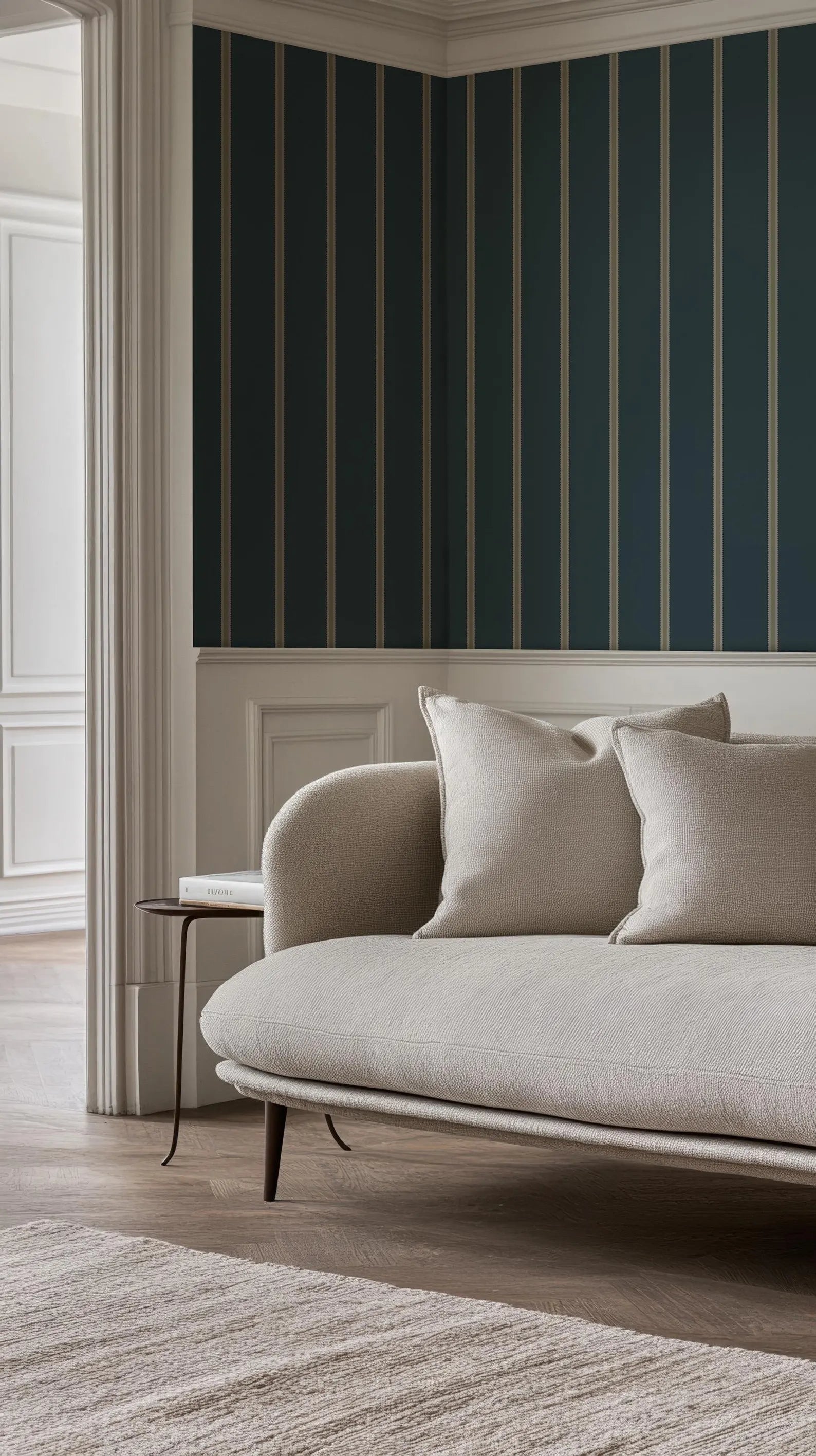 a modern living room with a white sofa and a wooden side table. The sofa has two pillows and is positioned against a wall with vertical stripes. The room has a wooden floor and a white rug, creating a clean and minimalist aesthetic.