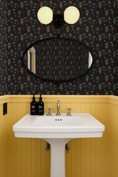a bathroom with a white pedestal sink, a mirror above it, and a floral wallpaper. There are two bottles of soap on the sink.