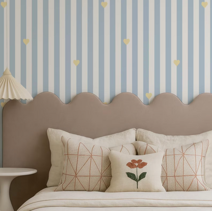 a cozy bedroom with a bed adorned with a wavy headboard and a single pillow featuring a flower design. The walls are decorated with blue and white striped wallpaper, and the room has a warm, inviting atmosphere.