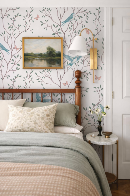 a cozy bedroom with a wooden headboard, a framed landscape painting, and a lamp on a side table. The room has a white and green color scheme, with a patterned wallpaper and a white bedspread.