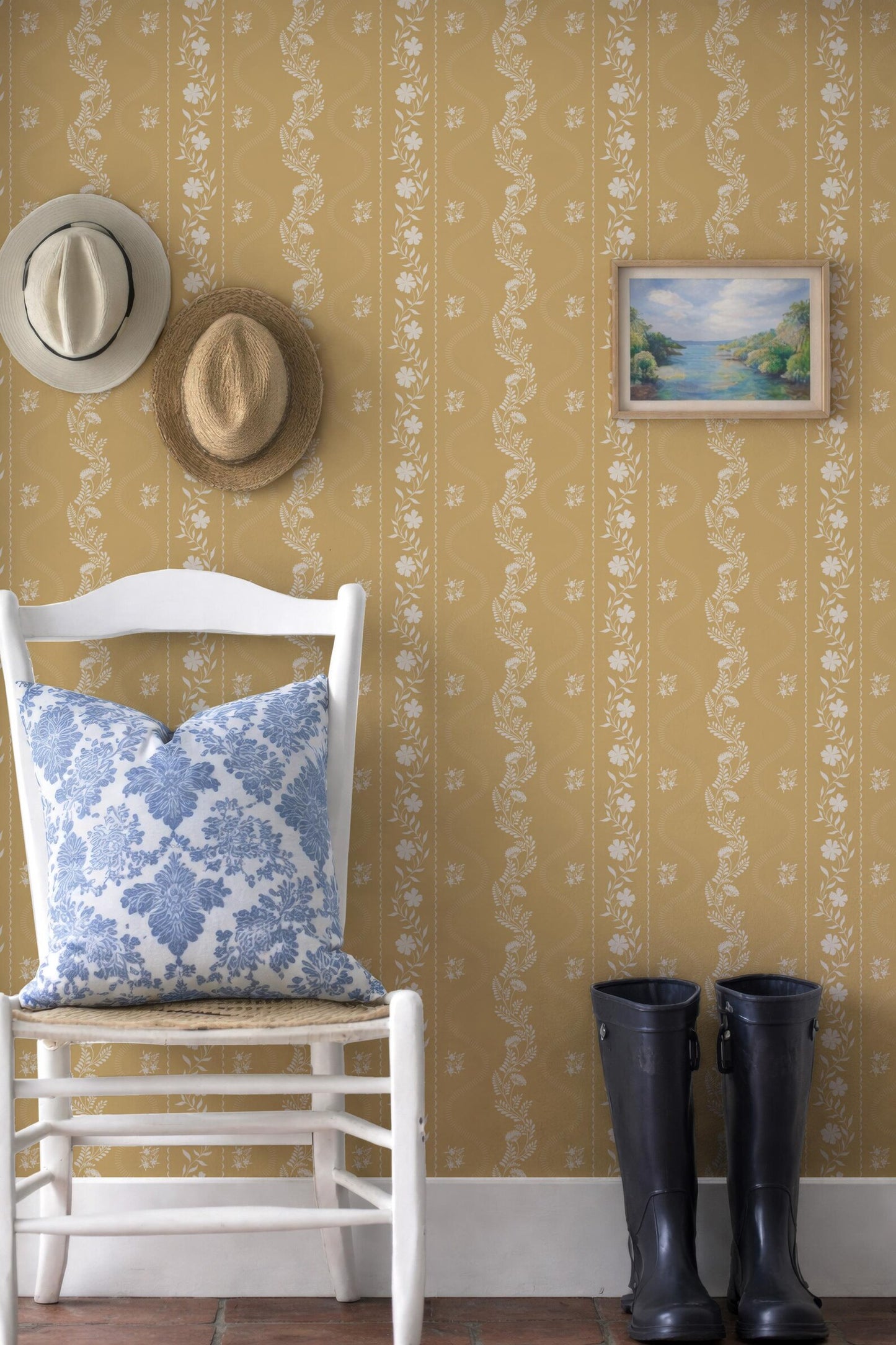 a cozy corner with a white chair, a hat, a painting, and a pair of black rain boots. The chair has a blue and white floral pillow on it. The wall behind the chair has a floral pattern, and there is a picture hanging on the wall.