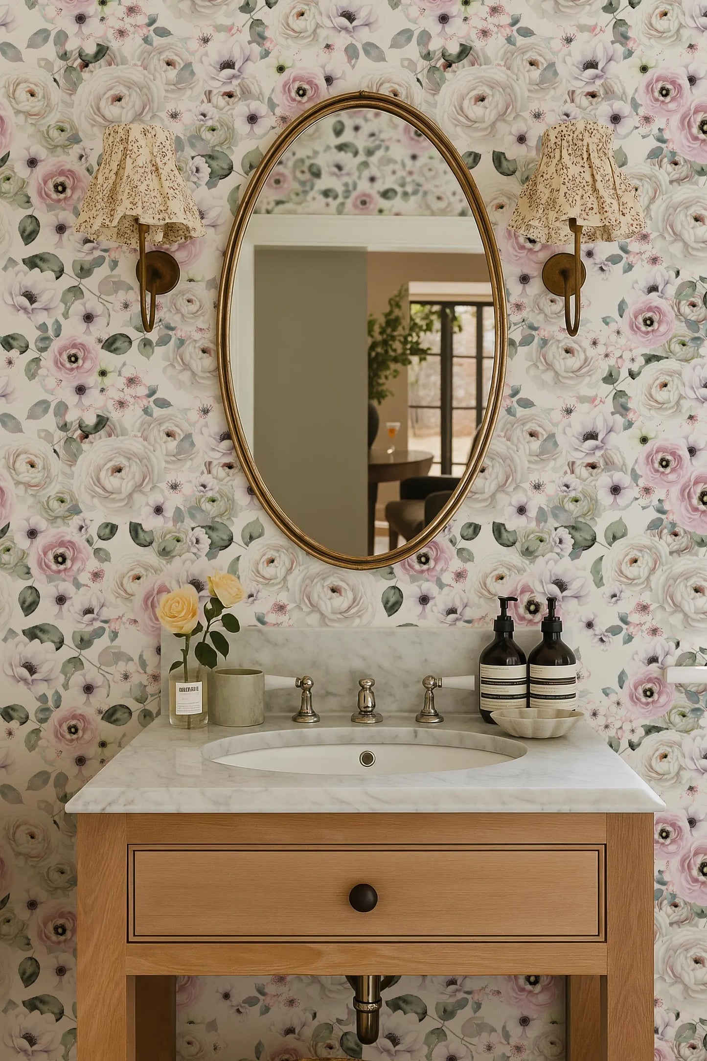 a bathroom with a white marble sink, a wooden vanity, and a large oval mirror with a gold frame. The walls are covered in a floral wallpaper, and there are two lamps on either side of the mirror. The sink is set into a wooden cabinet, and there are two bottles of soap on the counter.