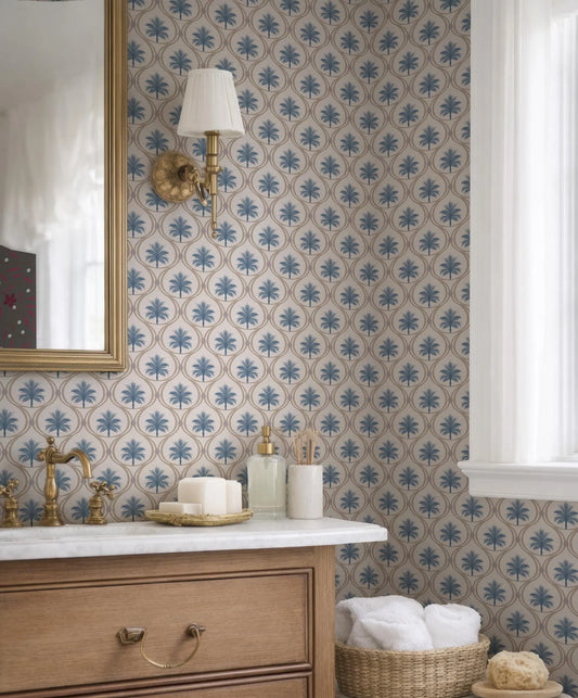 a bathroom with a white marble countertop, a gold faucet, and a mirror above it. The walls are covered in a blue and white patterned wallpaper, and there is a window with white curtains. On the countertop, there are various toiletries and towels, including a soap dispenser, a toothbrush holder, and a basket of folded towels.