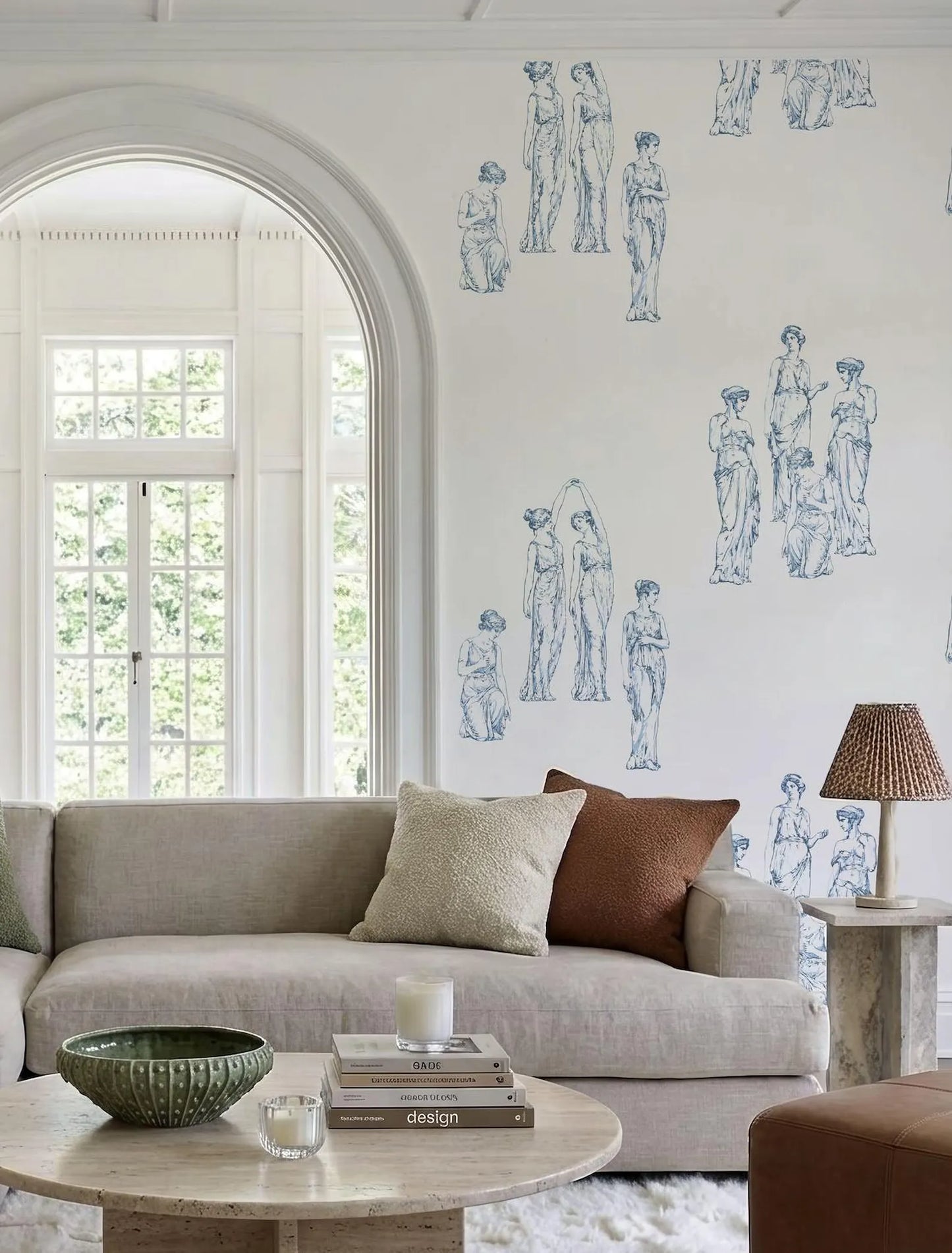 a cozy living room with a large arched window, a gray sofa, a round coffee table, and a lamp on a side table. The wall behind the sofa is decorated with blue and white illustrations of classical women.