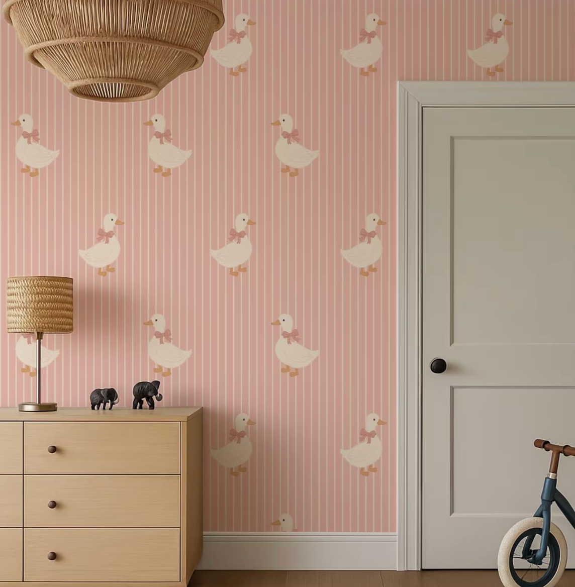 a pink wall with a repeating pattern of white ducks, a wooden dresser with a lamp, a bicycle, and a closed door.