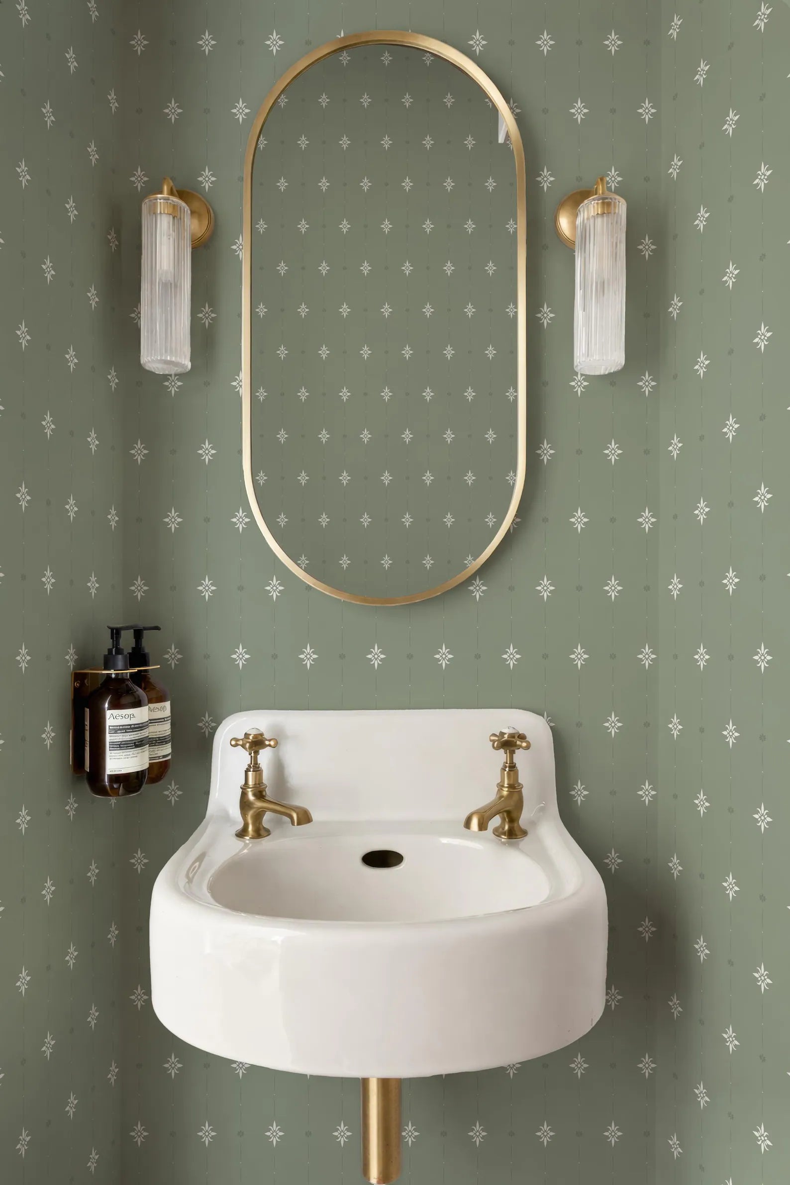 a bathroom with a white sink, a gold-framed mirror, and two clear glass lights on either side of the mirror. The walls are covered in a patterned green wallpaper with white stars.