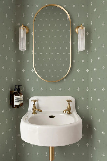 a bathroom with a white sink, a gold-framed mirror, and two clear glass lights on either side of the mirror. The walls are covered in a patterned green wallpaper with white stars.