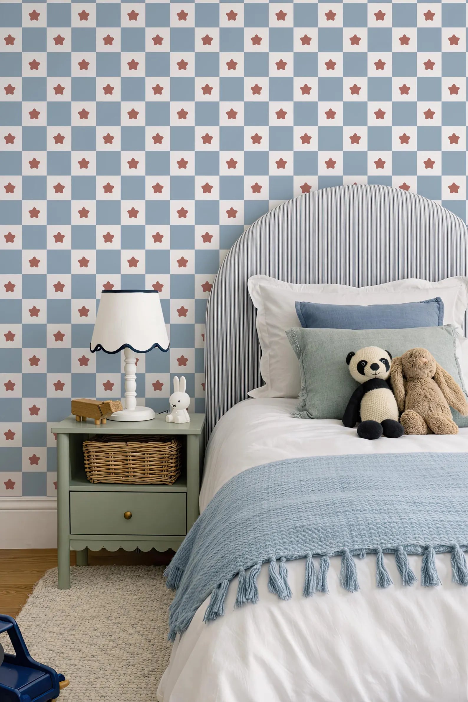 a cozy bedroom scene with a bed, nightstand, and stuffed animals. The bed has a striped headboard and is adorned with a blue blanket and a blue throw pillow. The nightstand has a white lamp and a small basket. A stuffed panda bear and a stuffed rabbit are resting on the bed. The wall behind the bed is decorated with a pattern of blue and white stars.