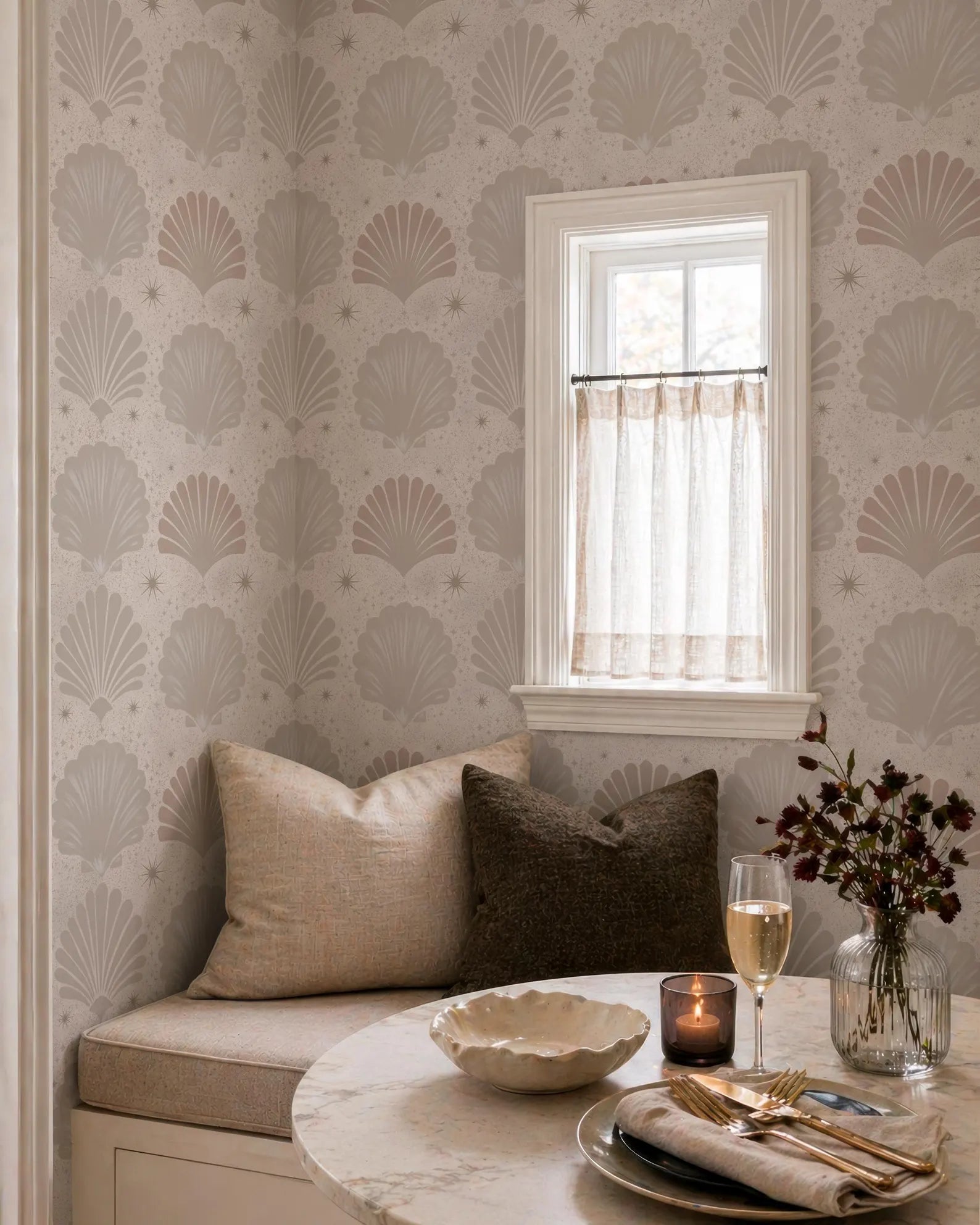 a cozy dining area with a round table set for two. The table is adorned with a vase of flowers, a candle, and a wine glass, creating a warm and inviting atmosphere. The table is surrounded by comfortable pillows and a window with a curtain, allowing natural light to illuminate the space. The overall scene conveys a sense of relaxation and enjoyment, with the carefully arranged table and decor suggesting a pleasant dining experience.