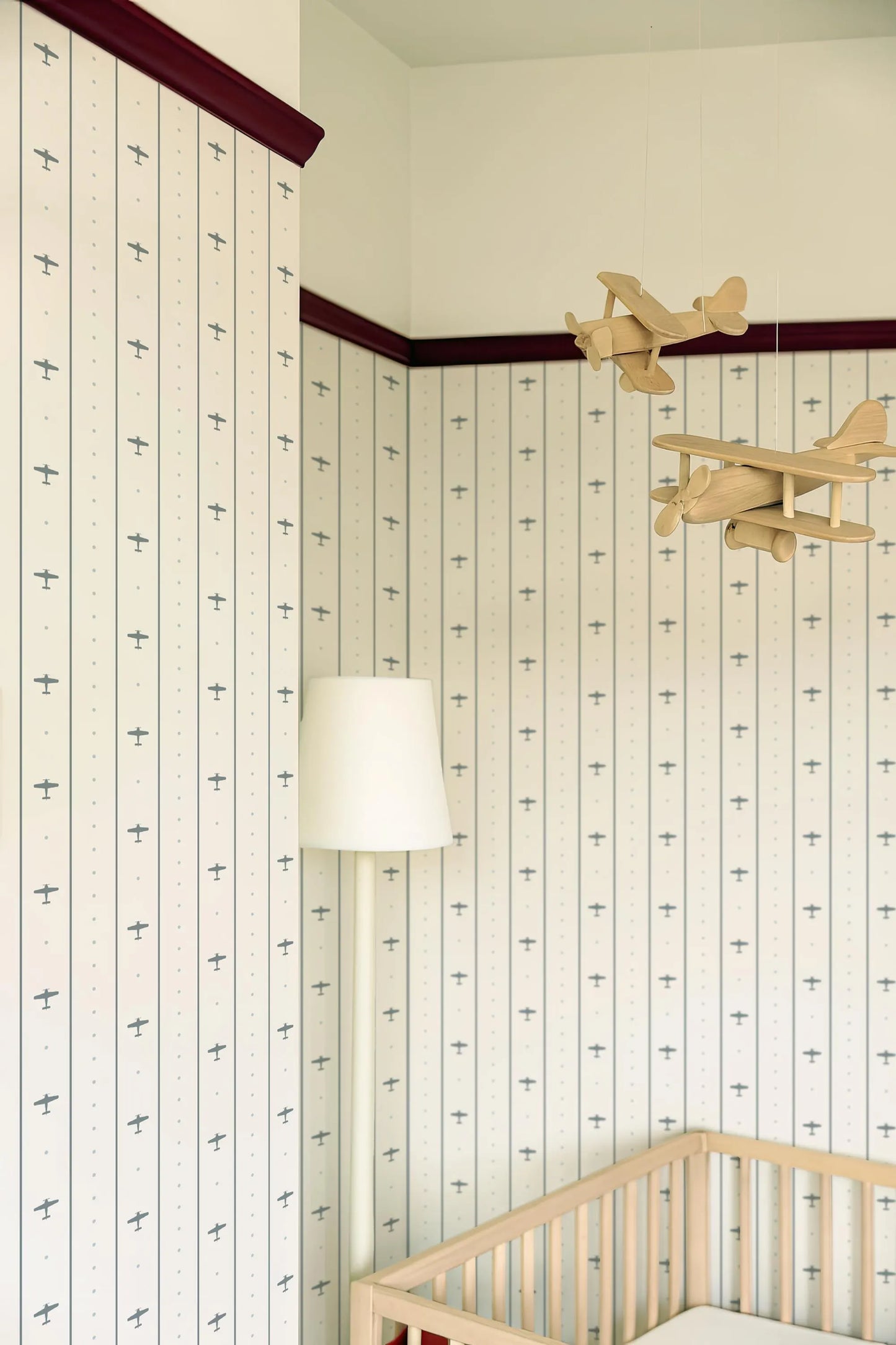 a nursery room with a wooden crib, a white lamp, and a wooden airplane hanging from the ceiling. The walls are decorated with a striped pattern, and the overall color scheme is predominantly white and blue.