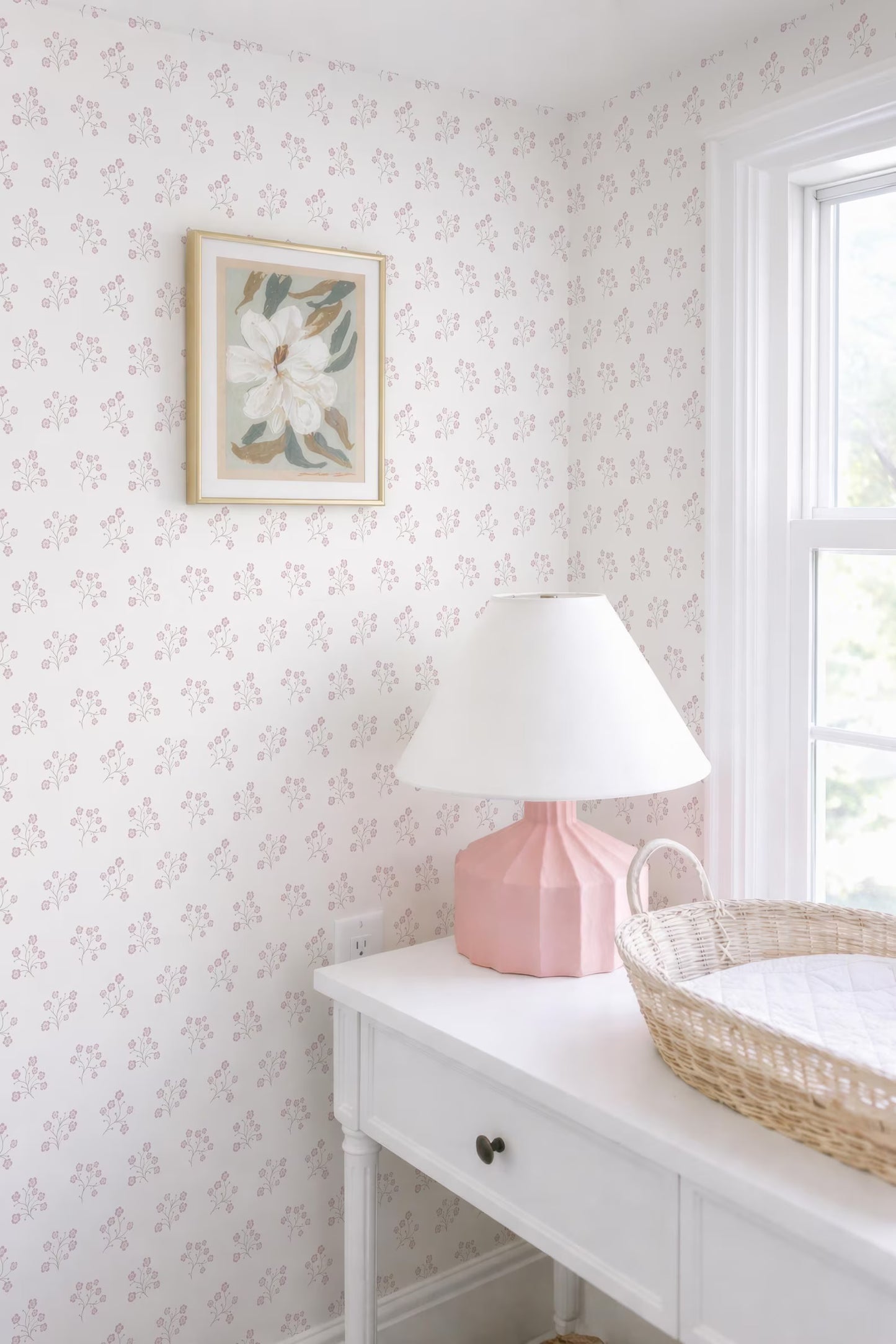 a white dresser with a pink lamp, a white picture frame, and a woven basket on top. The dresser is situated next to a window, and the wall behind it has a floral wallpaper.