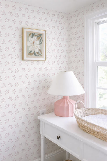 a white dresser with a pink lamp, a white picture frame, and a woven basket on top. The dresser is situated next to a window, and the wall behind it has a floral wallpaper.