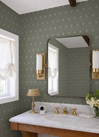 a bathroom with a white marble countertop, a gold-framed mirror, and a white lamp. The walls are covered in a green and white patterned wallpaper, and there are two white sconces on either side of the mirror. The bathroom also features a vase of white flowers on the countertop.