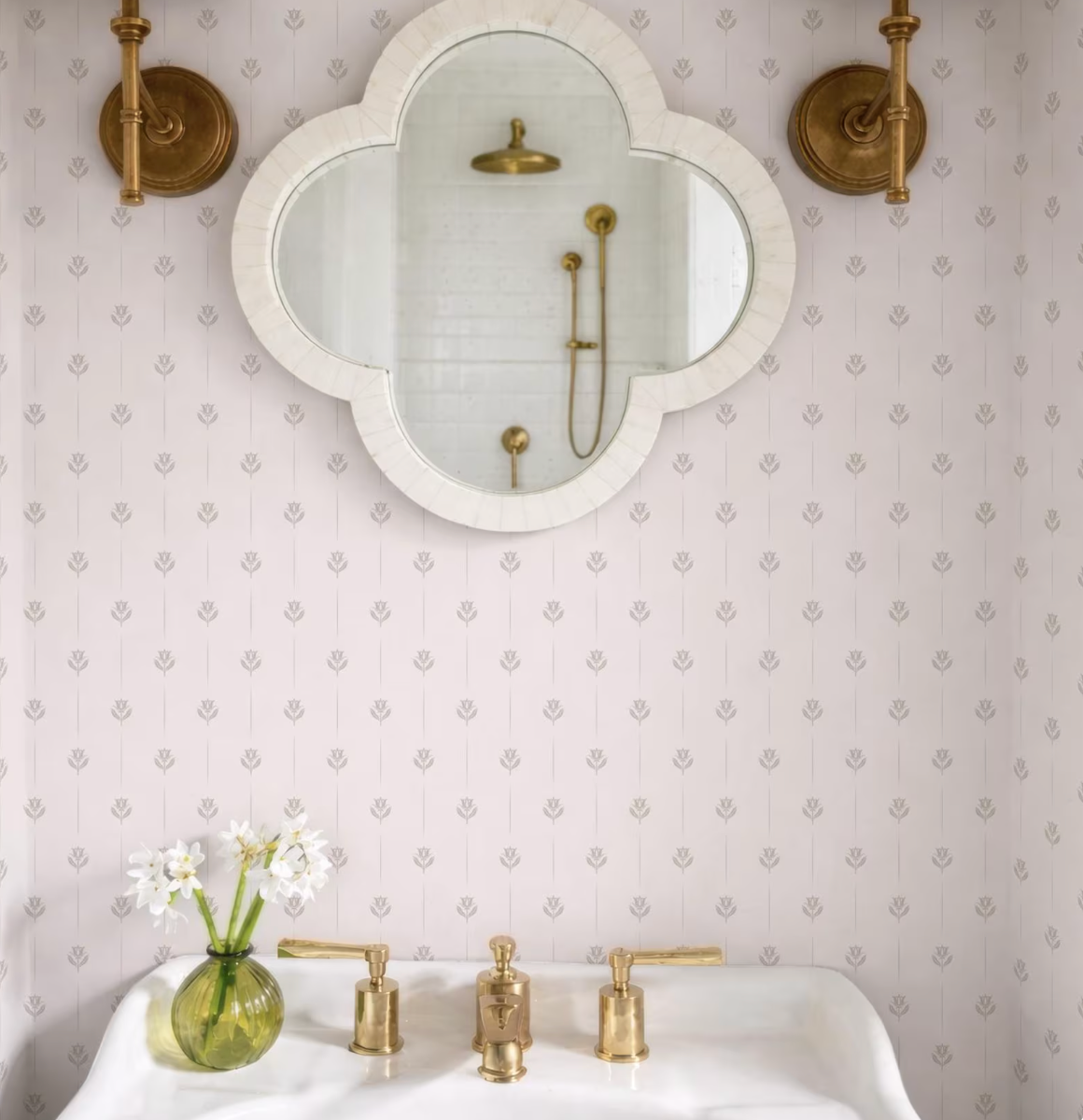a bathroom with a white sink, a mirror, and a vase of white flowers. The mirror is framed in a white oval shape and has a gold-colored frame. The wall behind the sink has a patterned wallpaper. The sink has a gold faucet and handles.