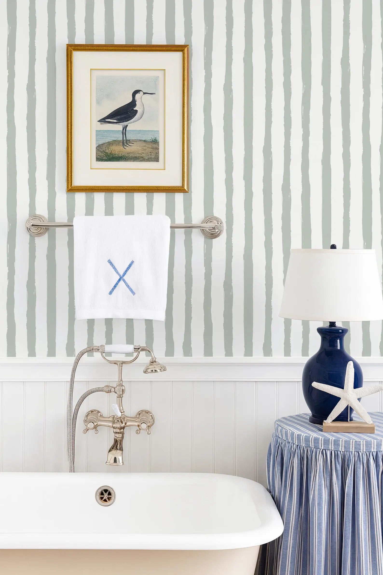 a bathroom with a white bathtub, a framed picture of a bird, a towel rack, a blue lamp, and a striped tablecloth.